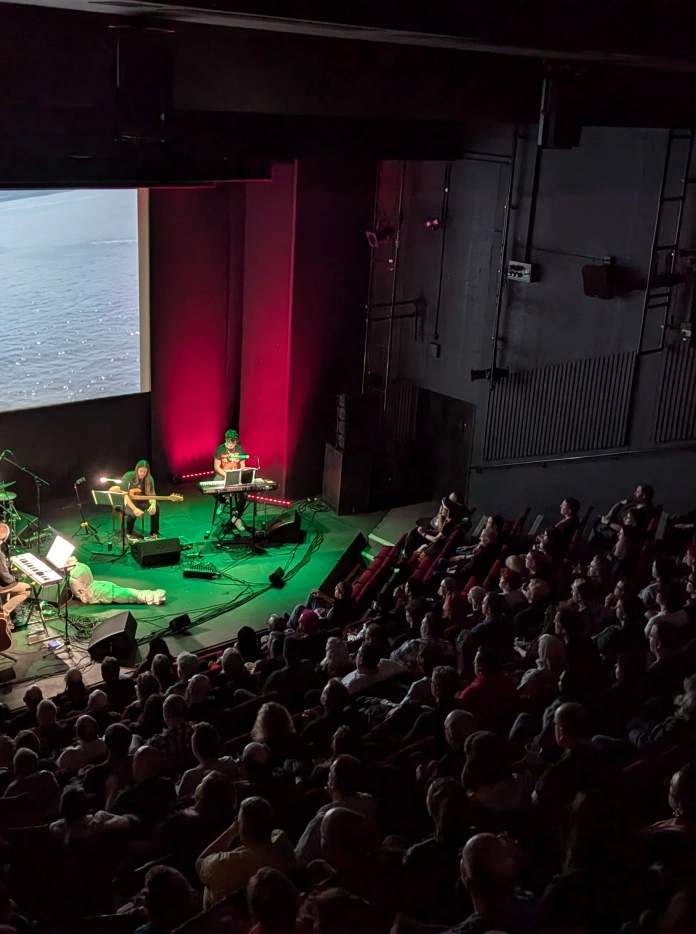 A live band performs on a stage beneath a large cinema screen showing a sailboat on water, with a packed audience watching on.